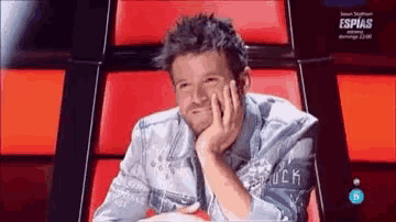 a man in a denim jacket is sitting in a red chair with his hand on his chin .