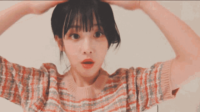 a woman wearing a pink and orange striped sweater has her hands on her head and a surprised look on her face