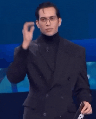 a man in a black suit and turtleneck is holding a cane while standing in front of a blue background .
