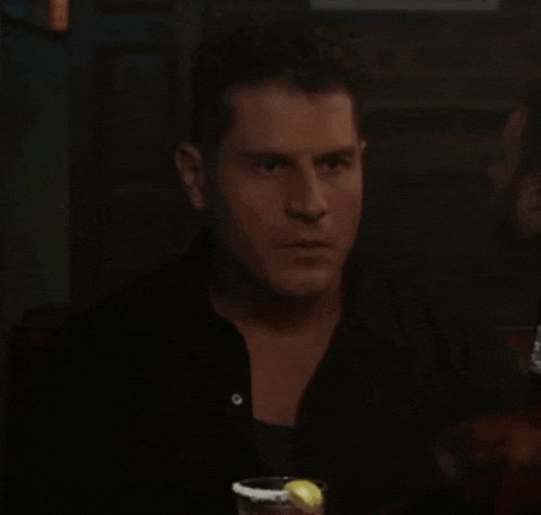 a man in a black shirt is sitting at a table with a drink in his hand .