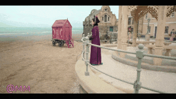 a woman in a purple coat is standing on a beach next to a fence with the letters @mm below her