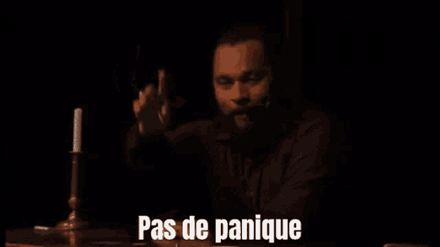 a man with a beard is sitting at a table with his hand to his mouth and the words pas de panique written above him