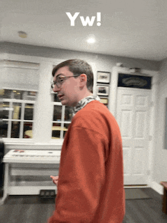 a man in an orange sweater is standing in a living room with a keyboard in the background and the words yw written on the bottom right