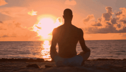 a man sits in a lotus position on the beach with the sun setting behind him