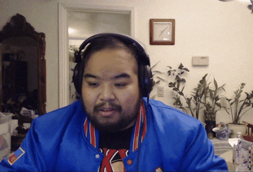 a man wearing headphones and a blue jacket looks at the camera with a clock on the wall behind him