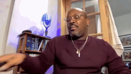 a man wearing glasses and a necklace is sitting in a chair in front of a microphone .