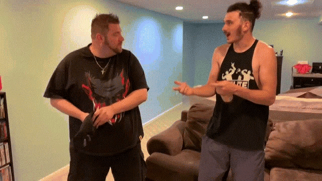 two men are standing in a living room and one of them is wearing a tank top that says ' elite ' on it