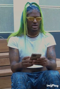 a man with green hair and yellow sunglasses is sitting on a bench looking at his phone