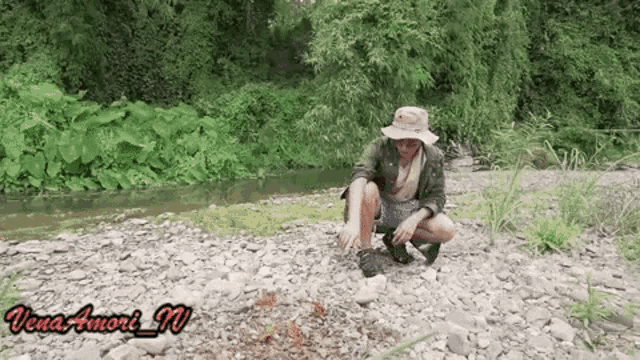 a man wearing a hat is kneeling on a rocky river bank .
