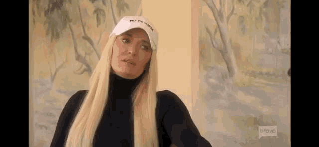 a blonde woman wearing a white hat and a black turtleneck is sitting in front of a painting .