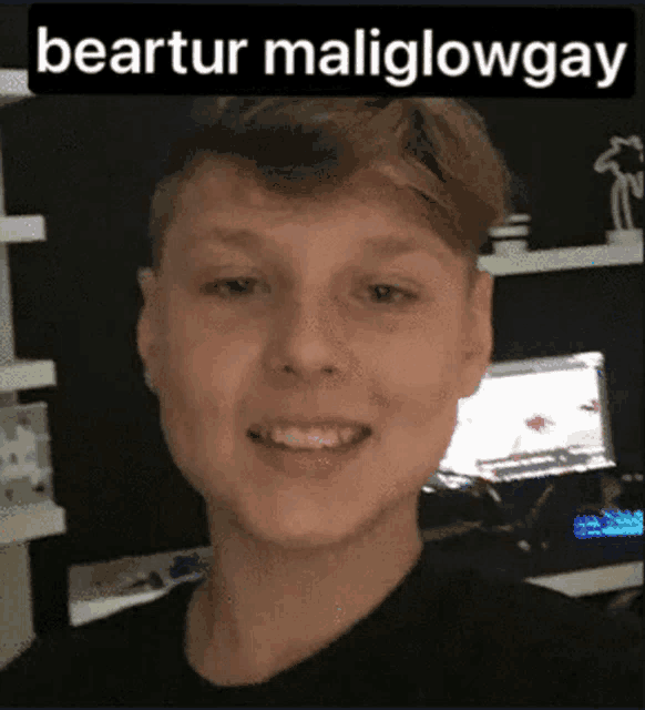 a young man in a black shirt is smiling in front of a computer screen with the words beartur maliglowgay written above him .