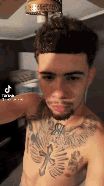 a shirtless man with a tattoo on his chest is taking a selfie with a ceiling fan in the background .