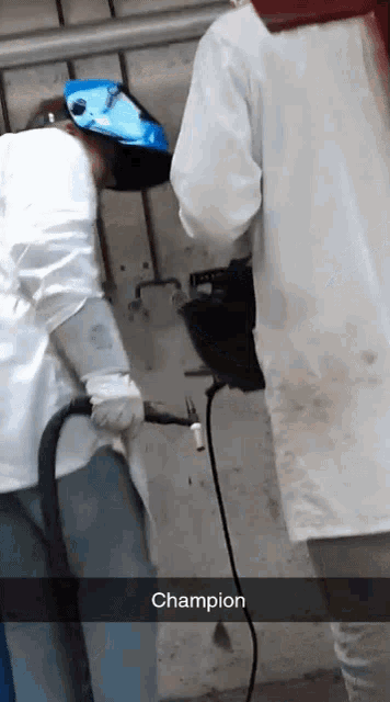 a man wearing a welding helmet is standing next to a man wearing a white coat with the word champion written on it