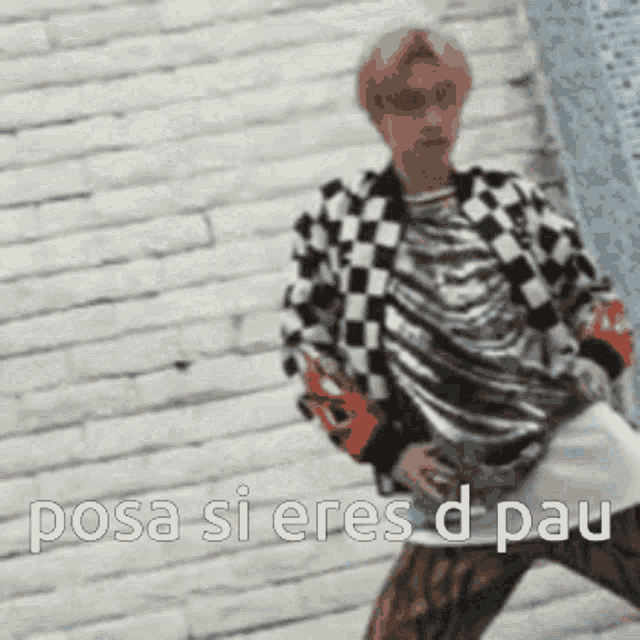 a man in a checkered jacket is standing in front of a brick wall with the words posa si eres d pau .