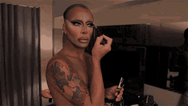 a shirtless drag queen is applying makeup to his face