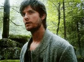 a man with a beard is standing in the woods wearing a sweater .
