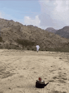 a man in a white robe is walking across a dirt field with mountains in the background