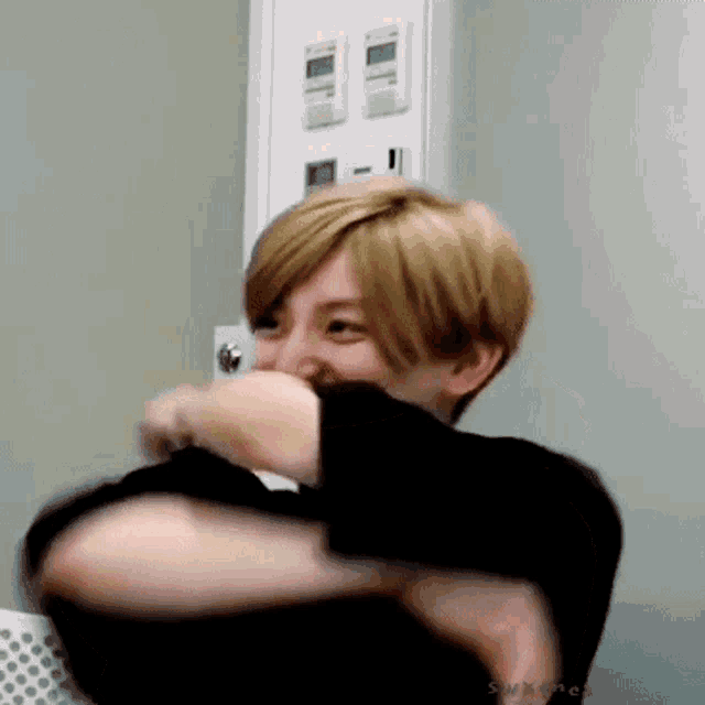a young man with blonde hair is laughing and covering his mouth with his hand while wearing a black shirt .
