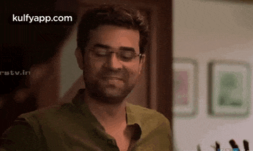 a man wearing glasses and a green shirt is smiling in a room with pictures on the wall .