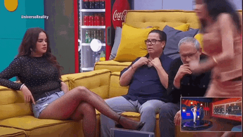a woman is sitting on a yellow couch next to a man and a woman with a coca cola cooler in the background
