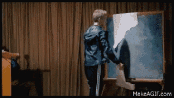 a man in a blue jacket is standing in front of a blackboard with makeagif.com written on it