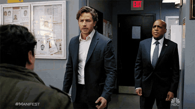 a man in a suit stands next to another man in a suit and tie in front of an exit sign