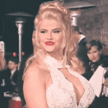 a woman in a white lace dress with a plunging neckline is standing in front of a group of people .