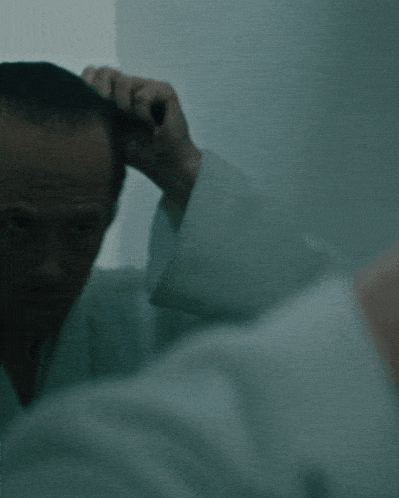 a man in a white shirt scratches his head in a dark room