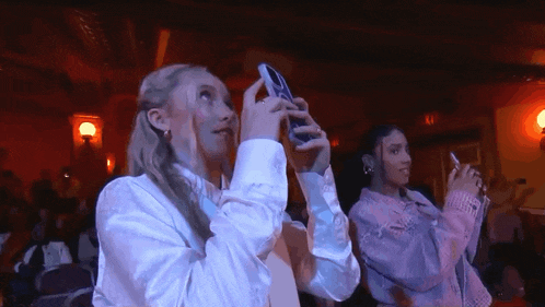 two women are taking a picture with their phones in a dark room .