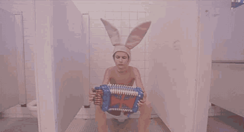 a person wearing bunny ears is holding an accordion in a bathroom stall