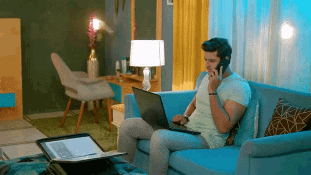 a man sitting on a couch talking on a cell phone while using a laptop