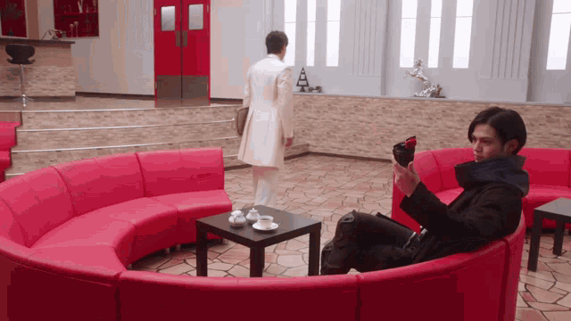 a man sits on a red couch in a living room while a man in a white coat stands behind him