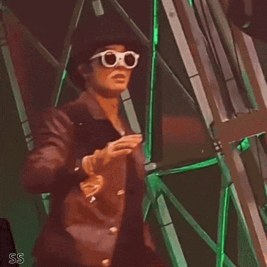 a man wearing sunglasses and a hat stands in front of a green wall