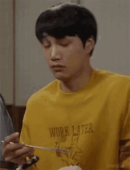 a young man wearing a yellow shirt that says `` work later '' is eating a spoonful of food .
