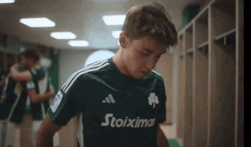 a man wearing a green adidas stoixima shirt stands in a locker room