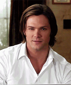 a man with long hair and a white shirt looks at the camera