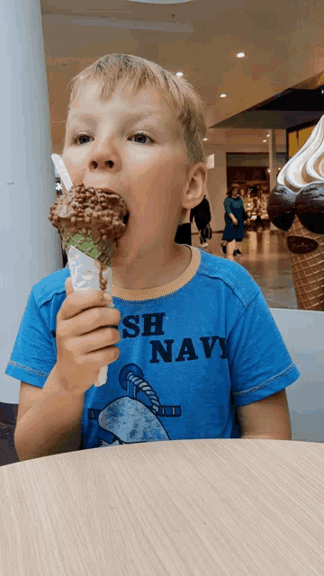 a young boy wearing a blue shirt that says navy is eating a chocolate ice cream cone