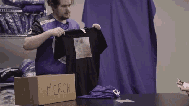 a man is holding a black shirt next to a box that says merch
