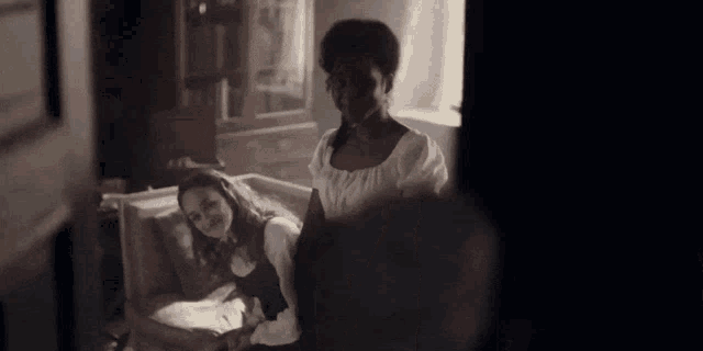 a woman is standing next to a woman sitting on a couch in a room .