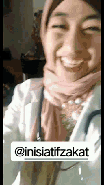 a woman wearing a pink scarf and a white coat is smiling with a sticker that says @inisiatifzakat