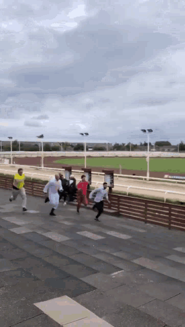 a group of people are running down a sidewalk in front of a race track