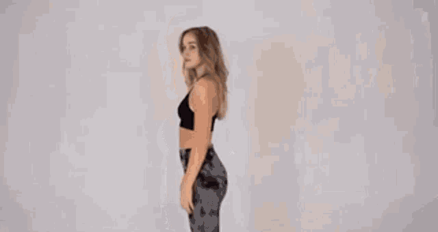 a woman in a black crop top and gray leggings is standing in front of a white wall .