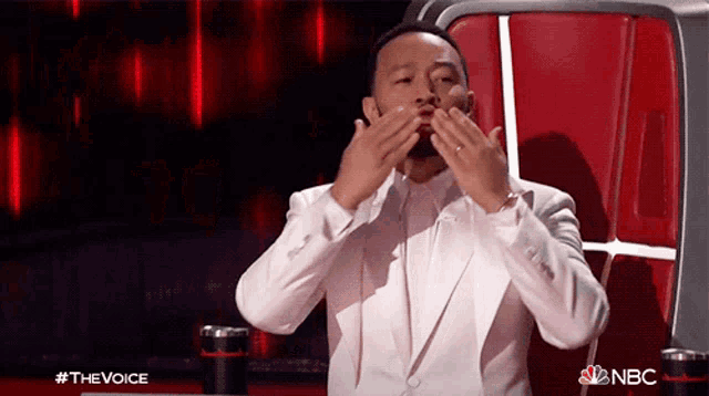 a man in a white suit is covering his mouth with his hands while sitting in front of a nbc logo