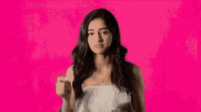 a young woman in a white dress is standing in front of a pink background and making a funny face .