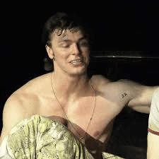 a shirtless man with a tattoo on his arm is sitting on a bed in a dark room .