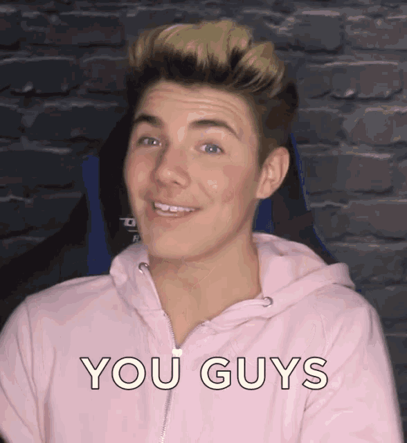 a man in a pink hoodie says you guys in front of a brick wall