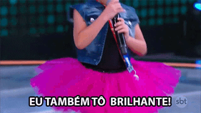 a girl in a pink tutu singing into a microphone with the words eu também to brilhante below her