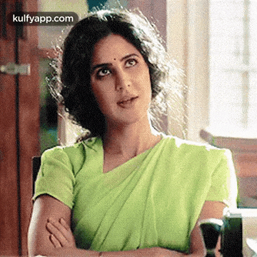 a woman in a green saree is sitting at a table with her arms crossed and looking up .