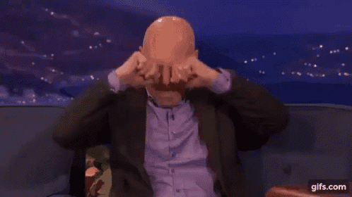 a bald man in a suit is sitting on a couch and covering his face with his hands .