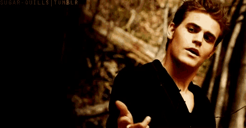 a man in a black shirt is standing in front of a stone wall with his hand out .